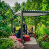 Eine Gruppe von Menschen unterhält sich unter einer manuellen Pergola mit Lamellendach in einem üppigen Garten. Sie sitzen um einen mit einer roten Tischdecke bedeckten Tisch inmitten von grünem Laub. Eine Person steht, während eine andere auf einem Korbstuhl sitzt. Auf dem Boden steht eine Laterne.