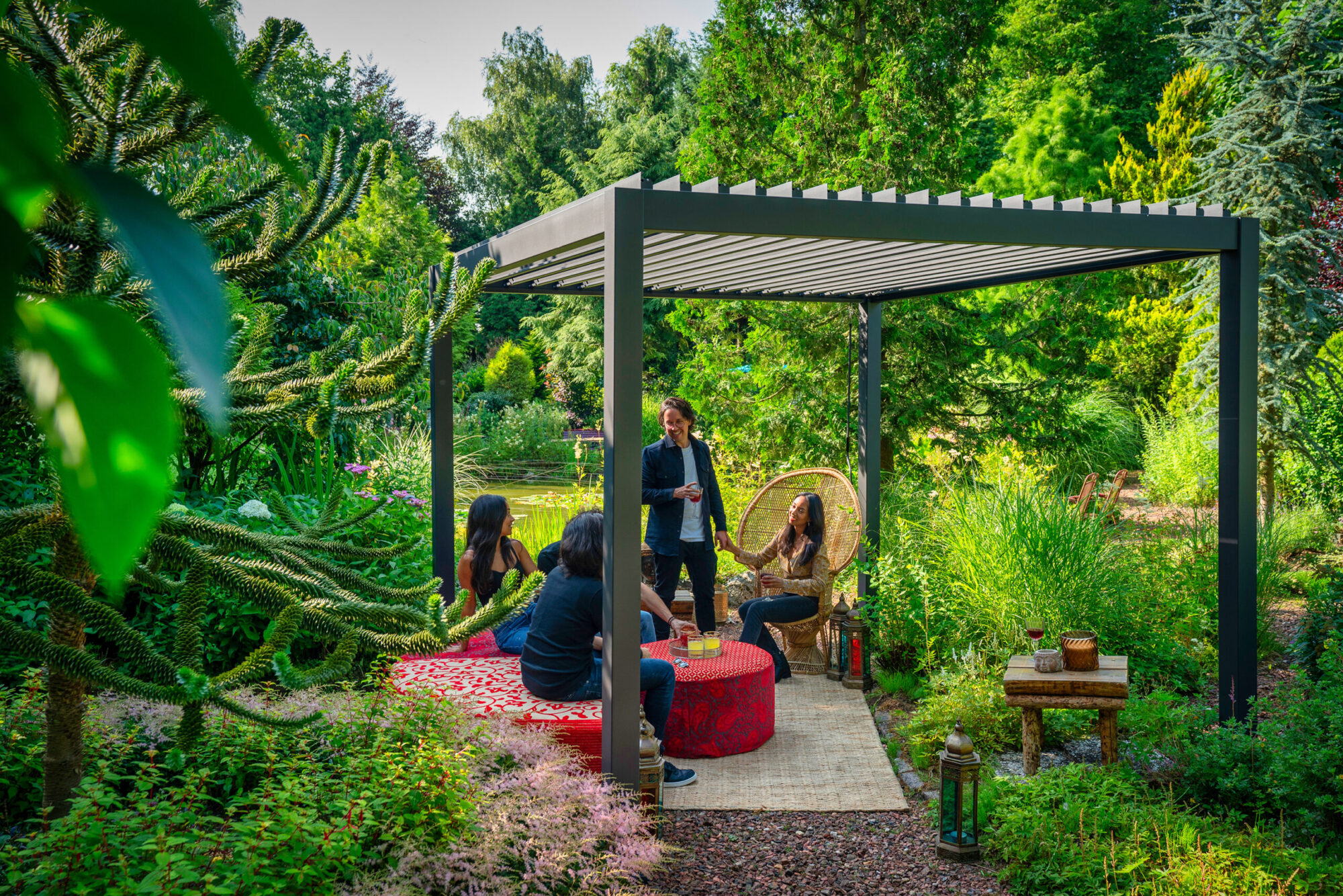 Eine Gruppe von Menschen unterhält sich unter einer manuellen Pergola mit Lamellendach in einem üppigen Garten. Sie sitzen um einen mit einer roten Tischdecke bedeckten Tisch inmitten von grünem Laub. Eine Person steht, während eine andere auf einem Korbstuhl sitzt. Auf dem Boden steht eine Laterne.