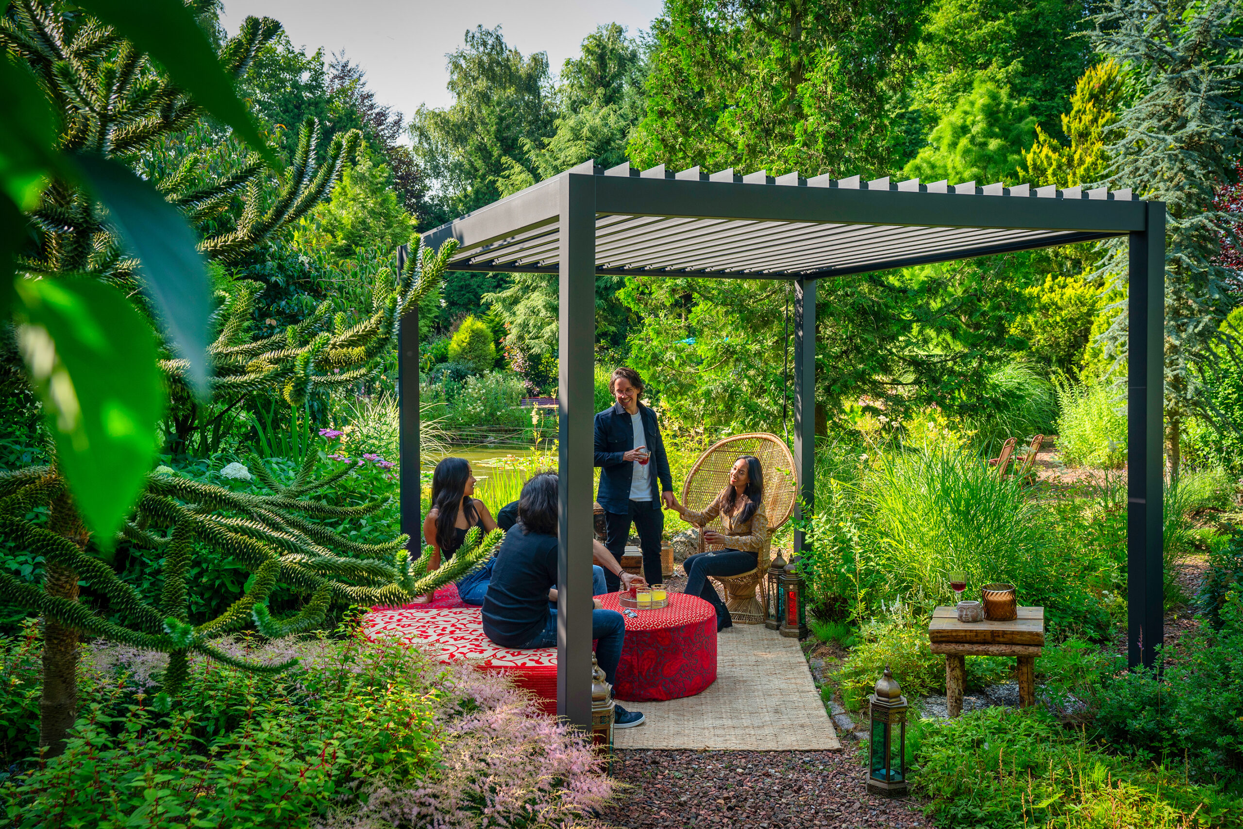 Eine Gruppe von Menschen unterhält sich unter einer manuellen Pergola mit Lamellendach in einem üppigen Garten. Sie sitzen um einen mit einer roten Tischdecke bedeckten Tisch inmitten von grünem Laub. Eine Person steht, während eine andere auf einem Korbstuhl sitzt. Auf dem Boden steht eine Laterne.