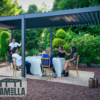 Eine Gruppe von Menschen speist im Freien unter einer dunkel gefärbten Pergola, aufgewertet durch das Lamellendach manuell, in einem üppigen Garten. Der Tisch ist mit weißen Tischdecken und Kerzen gedeckt. Einer der sechs Gäste steht auf, um anzustoßen. Die Umgebung strahlt eine entspannte, elegante Atmosphäre aus.