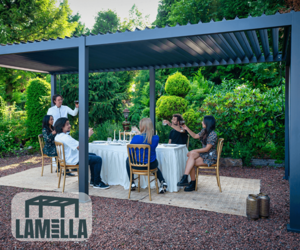 Eine Gruppe von Menschen speist im Freien unter einer dunkel gefärbten Pergola, aufgewertet durch das Lamellendach manuell, in einem üppigen Garten. Der Tisch ist mit weißen Tischdecken und Kerzen gedeckt. Einer der sechs Gäste steht auf, um anzustoßen. Die Umgebung strahlt eine entspannte, elegante Atmosphäre aus.