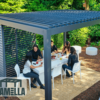 Fünf Personen versammeln sich um einen weißen Tisch unter einer eleganten, modernen Pergola mit Lamellendach in einem üppigen Garten. Sie genießen Getränke und Snacks umgeben von farbenfrohen Pflanzen und Sträuchern. Das „Lamella“-Logo ist dezent in der Ecke platziert.