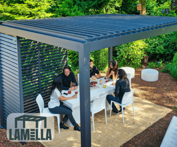 Fünf Personen versammeln sich um einen weißen Tisch unter einer eleganten, modernen Pergola mit Lamellendach in einem üppigen Garten. Sie genießen Getränke und Snacks umgeben von farbenfrohen Pflanzen und Sträuchern. Das „Lamella“-Logo ist dezent in der Ecke platziert.