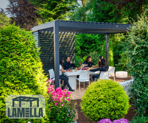 Eine Gruppe von Menschen sitzt um einen weißen Tisch und genießt eine Mahlzeit unter dem stilvollen schwarzen Lamellendach in einem üppigen Garten. Der Bereich ist von leuchtend grünen Pflanzen und bunten Blumen umgeben, und in der unteren linken Ecke erscheint das Lamella-Logo.