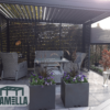 Eine moderne Außenterrasse verfügt über eine Pergola mit Korbmöbeln wie einem Sofa und Stühlen um einen Glastisch. Unter dem manuell bedienbaren Lamellendach stehen davor zwei Pflanzkästen mit violetten Blumen und Grünpflanzen, während Lamellenwände den Raum teilweise abgrenzen.