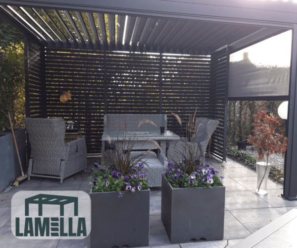 Eine moderne Außenterrasse verfügt über eine Pergola mit Korbmöbeln wie einem Sofa und Stühlen um einen Glastisch. Unter dem manuell bedienbaren Lamellendach stehen davor zwei Pflanzkästen mit violetten Blumen und Grünpflanzen, während Lamellenwände den Raum teilweise abgrenzen.