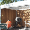 Eine Außenterrasse mit einer hölzernen Sichtschutzwand präsentiert einen roten Keramikgrill und einen holzbefeuerten Pizzaofen. Eine manuelle Lamellendach-Pergola spendet Schatten und ergänzt den kleinen Essbereich mit weißen Stühlen. Das LAMELLA-Logo ist in der Ecke prominent angebracht.