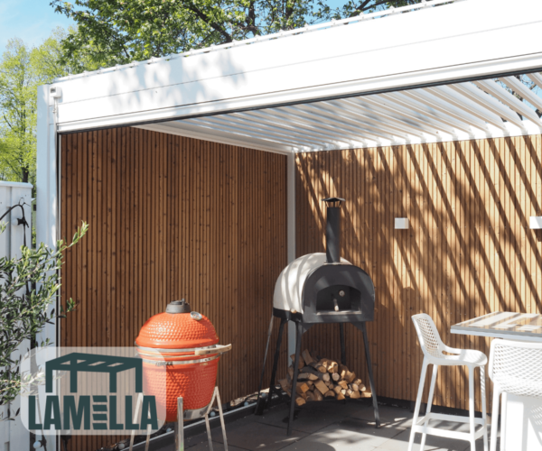 Eine Außenterrasse mit einer hölzernen Sichtschutzwand präsentiert einen roten Keramikgrill und einen holzbefeuerten Pizzaofen. Eine manuelle Lamellendach-Pergola spendet Schatten und ergänzt den kleinen Essbereich mit weißen Stühlen. Das LAMELLA-Logo ist in der Ecke prominent angebracht.