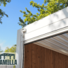 Eine moderne Pergola mit dem manuellen Lamellendach, einem einziehbaren Dach, ist an einem sonnigen Tag zu sehen. Die Struktur hat einen makellos weißen Rahmen und holzgetäfelte Seiten, mit Bäumen aus grünen Blättern, die den strahlend blauen Himmel umrahmen. In der unteren linken Ecke ist das LAMELLA-Logo deutlich sichtbar.