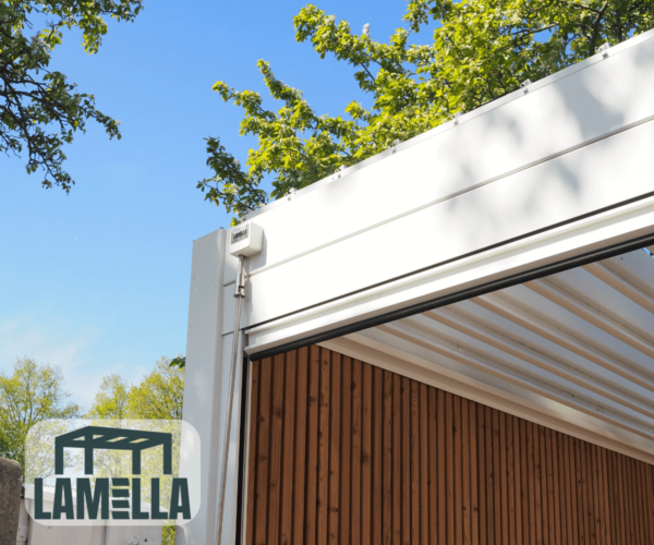 Eine moderne Pergola mit dem manuellen Lamellendach, einem einziehbaren Dach, ist an einem sonnigen Tag zu sehen. Die Struktur hat einen makellos weißen Rahmen und holzgetäfelte Seiten, mit Bäumen aus grünen Blättern, die den strahlend blauen Himmel umrahmen. In der unteren linken Ecke ist das LAMELLA-Logo deutlich sichtbar.