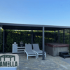 Eine moderne Außenterrasse präsentiert eine stilvolle Lamellendach-Pergola in Schwarz, begleitet von zwei bequemen Loungesesseln, einem integrierten Grill und einem eleganten Whirlpool. Der Steinboden ist elegant von leuchtend grünen Hecken gesäumt. In der unteren linken Ecke befindet sich ein Logo mit der Aufschrift „LAMELLA“.