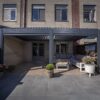 Eine stilvolle überdachte Terrasse mit Sitzgelegenheiten im Freien, Topfpflanzen und einem Couchtisch umfasst das manuell verstellbare Lamellendach, das an ein Backsteinhaus mit großen Fenstern und Doppeltüren zum Innenraum angeschlossen ist.