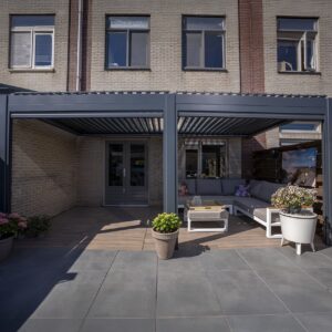 Eine stilvolle überdachte Terrasse mit Sitzgelegenheiten im Freien, Topfpflanzen und einem Couchtisch umfasst das manuell verstellbare Lamellendach, das an ein Backsteinhaus mit großen Fenstern und Doppeltüren zum Innenraum angeschlossen ist.