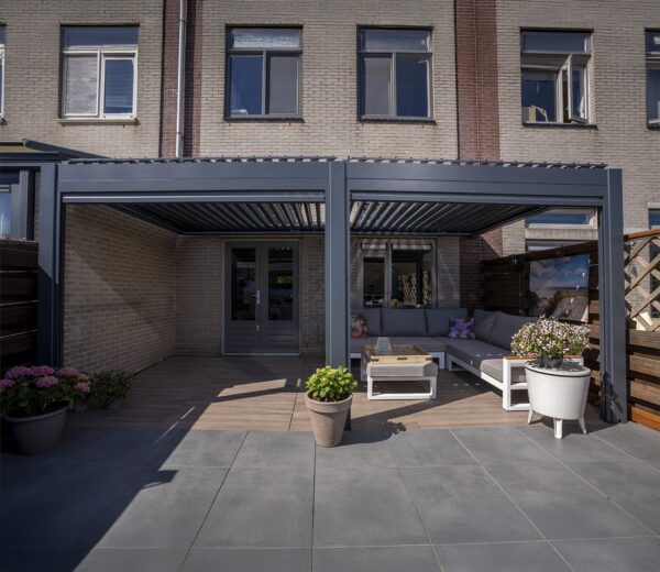 Eine stilvolle überdachte Terrasse mit Sitzgelegenheiten im Freien, Topfpflanzen und einem Couchtisch umfasst das manuell verstellbare Lamellendach, das an ein Backsteinhaus mit großen Fenstern und Doppeltüren zum Innenraum angeschlossen ist.