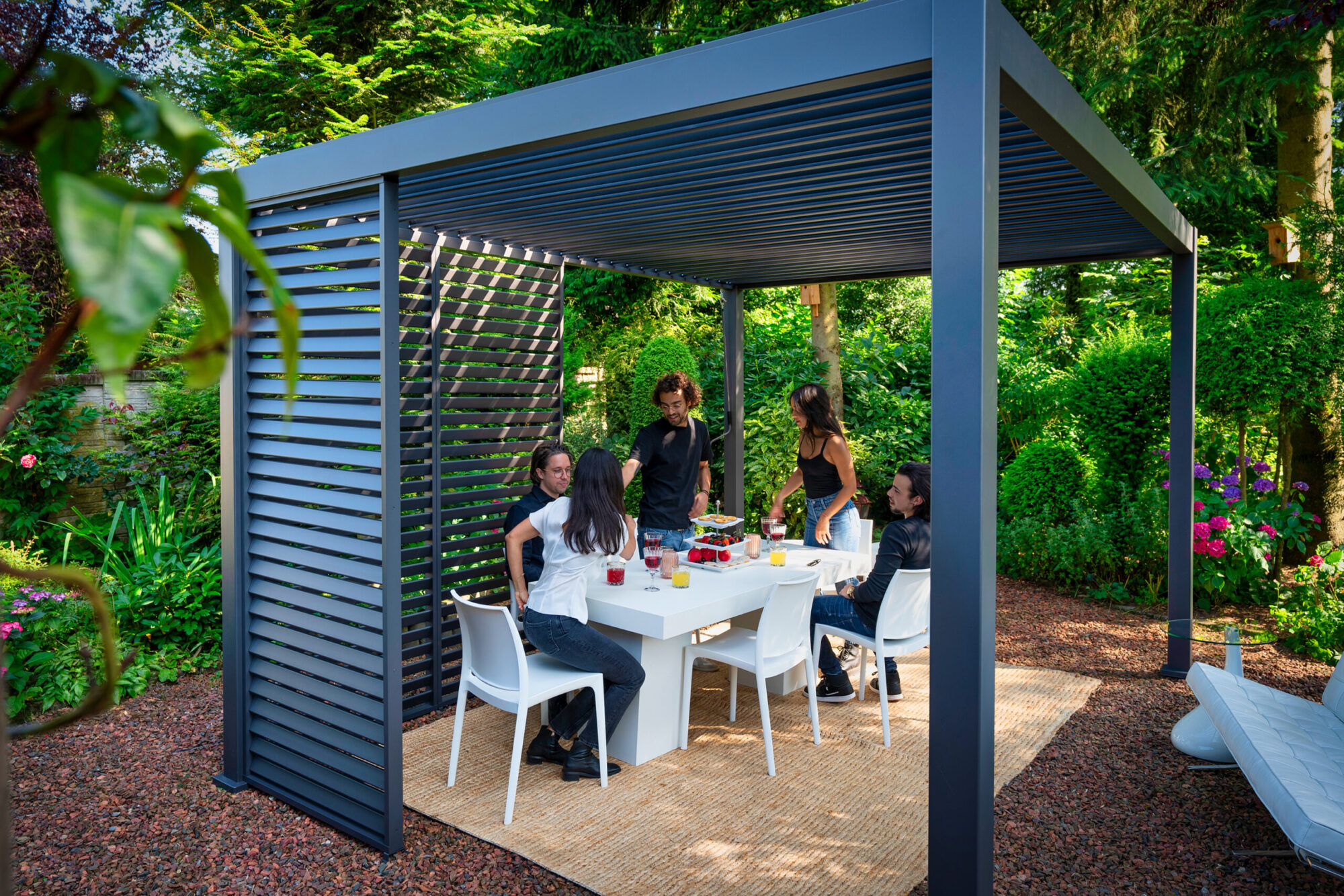 Eine Gruppe genießt Essen und Getränke an einem Tisch unter einem maßgefertigten Lamellendach nach Maß (Preis auf Anfrage) in einem grünen Garten, umgeben von grünen Pflanzen und Bäumen. Der Boden ist mit Kies bedeckt, hervorgehoben durch einen gewebten Teppich. Die Atmosphäre ist hell und fröhlich.