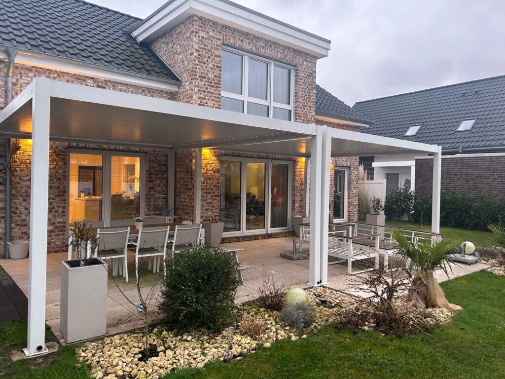 Ein modernes Backsteinhaus mit einer geräumigen Terrasse verfügt über eine weiße Pergola mit Lamellendach und elektrischem Antrieb. Darunter befindet sich der Außenbereich mit einem Esstisch und Stühlen. Der Garten ist mit Steinen, Sträuchern und Zierpflanzen angelegt.