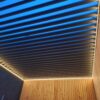 Eine Pergola mit LED-Beleuchtung und Lamellendach verfügt über horizontale Lamellen mit warmer Beleuchtung an den Rändern. Die Wände bestehen aus vertikalen Holzbrettern und die offenen Lamellen geben den Blick auf den Himmel frei und lassen erahnen, ob es Morgen- oder Abenddämmerung ist.