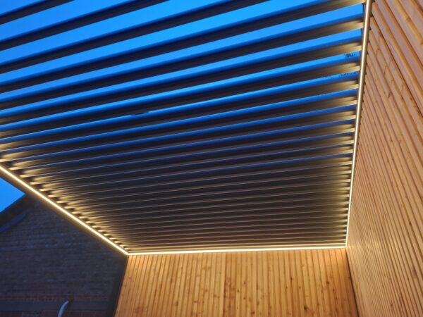 Eine Pergola mit LED-Beleuchtung und Lamellendach verfügt über horizontale Lamellen mit warmer Beleuchtung an den Rändern. Die Wände bestehen aus vertikalen Holzbrettern und die offenen Lamellen geben den Blick auf den Himmel frei und lassen erahnen, ob es Morgen- oder Abenddämmerung ist.