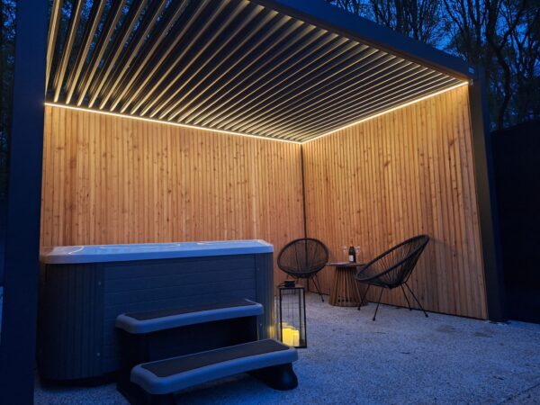 In der Abenddämmerung lädt der Außenbereich mit einem modernen Whirlpool und Treppen unter einer Pergola mit LED-Beleuchtung und Lamellendach zum Verweilen ein. Zwei dunkle Stühle, ein kleiner Tisch mit Getränken und eine Laterne mit Kerzen stehen auf dem Kiesboden, während Bäume eine ruhige Kulisse bieten.
