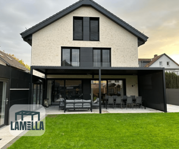 Ein modernes zweistöckiges Haus verfügt über eine beige Ziegelfassade mit schwarzen Fensterläden. Eine Pergola mit dem Lamellendach-Elektrisch-System spendet Schatten auf einer Terrasse mit Sitzgelegenheiten im Freien, die auf einen üppigen grünen Rasen hinausführt. An der Ecke ist das unverwechselbare Lamella-Logo zu sehen.