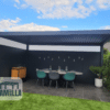 Eine moderne Außenterrasse präsentiert eine elegante Pergola mit Lamellendach Elektrisch, dazu einen Esstisch und Stühle auf gefliestem Boden. Links stehen ein Schrank und ein Grill, während eine Topfpflanze die rechte Seite schmückt. Die Szene spielt vor einem Hintergrund aus blauem Himmel mit Wolken. In der unteren linken Ecke befindet sich das LAMELLA-Logo.