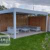 Eine moderne Pergola mit weißem Metallrahmen und Holzlamellenwänden präsentiert das elegante Design von Lamellendach Elektrisch. Sie umfasst weiße Lamellensitze und einen zentralen Couchtisch auf einer gepflasterten Terrasse, umgeben von Gras und üppigem Grün, alles unter einem bewölkten Himmel. Das LAMELLA-Logo unterstreicht den innovativen Stil.