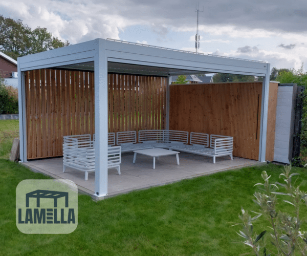 Eine moderne Pergola mit weißem Metallrahmen und Holzlamellenwänden präsentiert das elegante Design von Lamellendach Elektrisch. Sie umfasst weiße Lamellensitze und einen zentralen Couchtisch auf einer gepflasterten Terrasse, umgeben von Gras und üppigem Grün, alles unter einem bewölkten Himmel. Das LAMELLA-Logo unterstreicht den innovativen Stil.