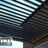 Ein Blick nach oben auf eine elegante, moderne Pergola, die an ein raffiniertes Lamellendach nach Maß erinnert, mit gleichmäßig angeordneten dunklen Metalllamellen vor einem klaren blauen Himmel. Ein Teil eines Backsteingebäudes ist sichtbar, während in der unteren linken Ecke das Lamella-Logo zu sehen ist.