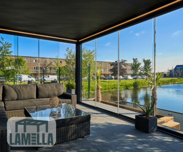 Eine moderne Außenterrasse mit Glaswänden und einem eleganten Lamellendach mit Blick auf einen Teich und Gebäude. Sie verfügt über Korbmöbel mit dunklen Kissen, einen reflektierenden Couchtisch und Topfpflanzen. Mit einem LAMELLA-Logo in der unteren linken Ecke versehen.