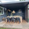 Ein gemütlicher Essbereich im Freien verfügt über einen Holztisch und schwarze Stühle unter einer eleganten Pergola mit Lamellen. Zur Einrichtung gehören eine große Hängelampe aus Korbgeflecht, Wandpflanzen und gerahmte Kunstwerke. Der geometrisch gemusterte Boden wird für zusätzlichen Charme von einer stilvollen Lamellenschiebewand oder einem festen Lamellenpaneel eingefasst.