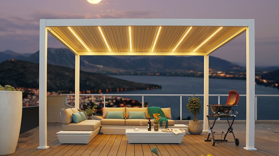 Eine moderne Dachterrasse mit einem maßgeschneiderten LED-Lamellendach nach Maß wirft in der Abenddämmerung ein warmes Licht. Entspannen Sie auf dem gemütlichen Sofa mit Kissen neben dem Couchtisch und Grill und genießen Sie den ruhigen Blick auf die Berge und den See bei Vollmond.