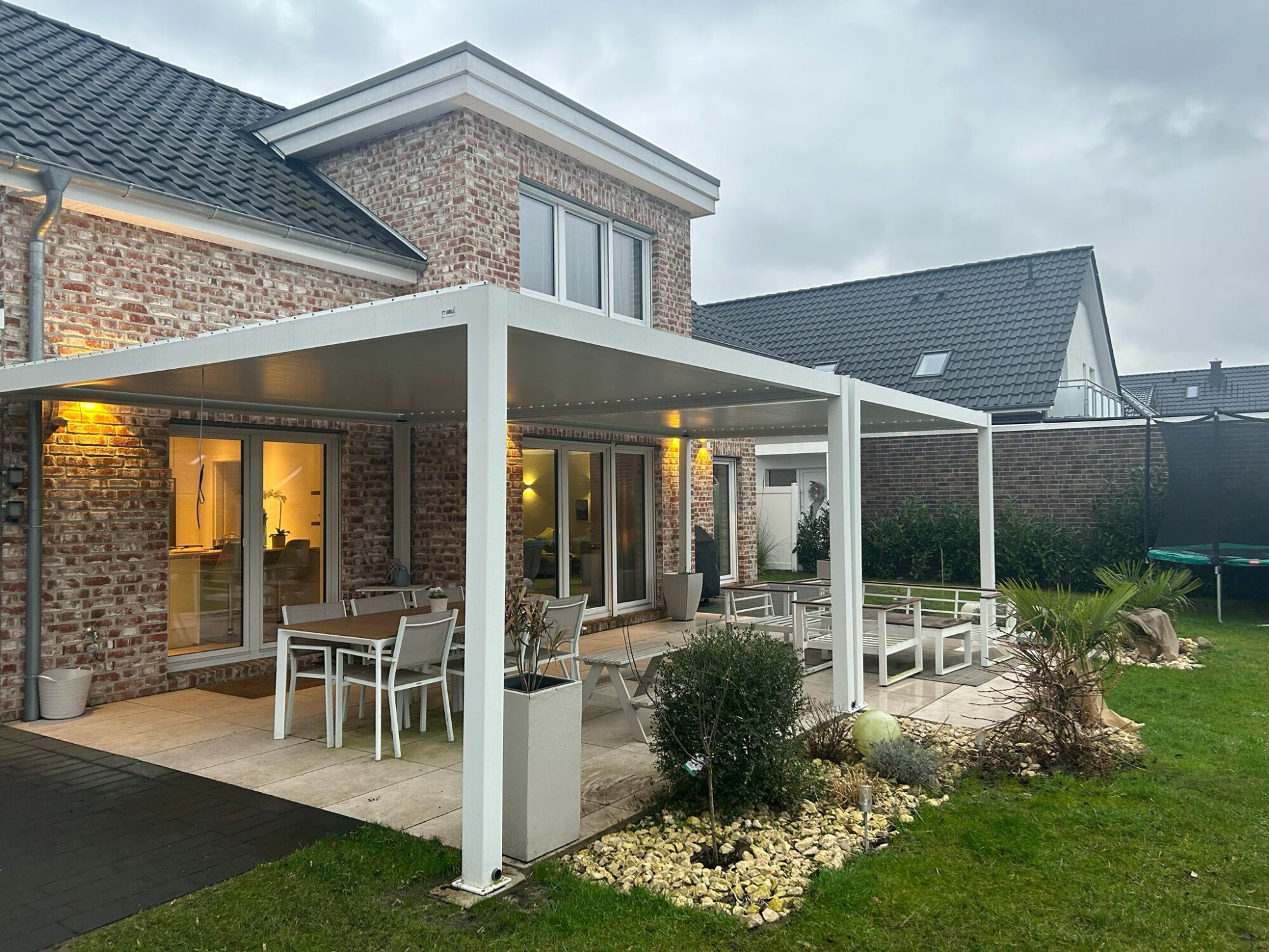 Ein modernes Backsteinhaus mit großen Fenstern und einer überdachten Terrasse mit Esstischen und Stühlen im Freien unter einer maßgeschneiderten Überdachung aus Aluminiumlamellen. Der Hof ist mit Steinen, Pflanzen und einem kleinen Trampolin angelegt. Der Himmel ist bedeckt.