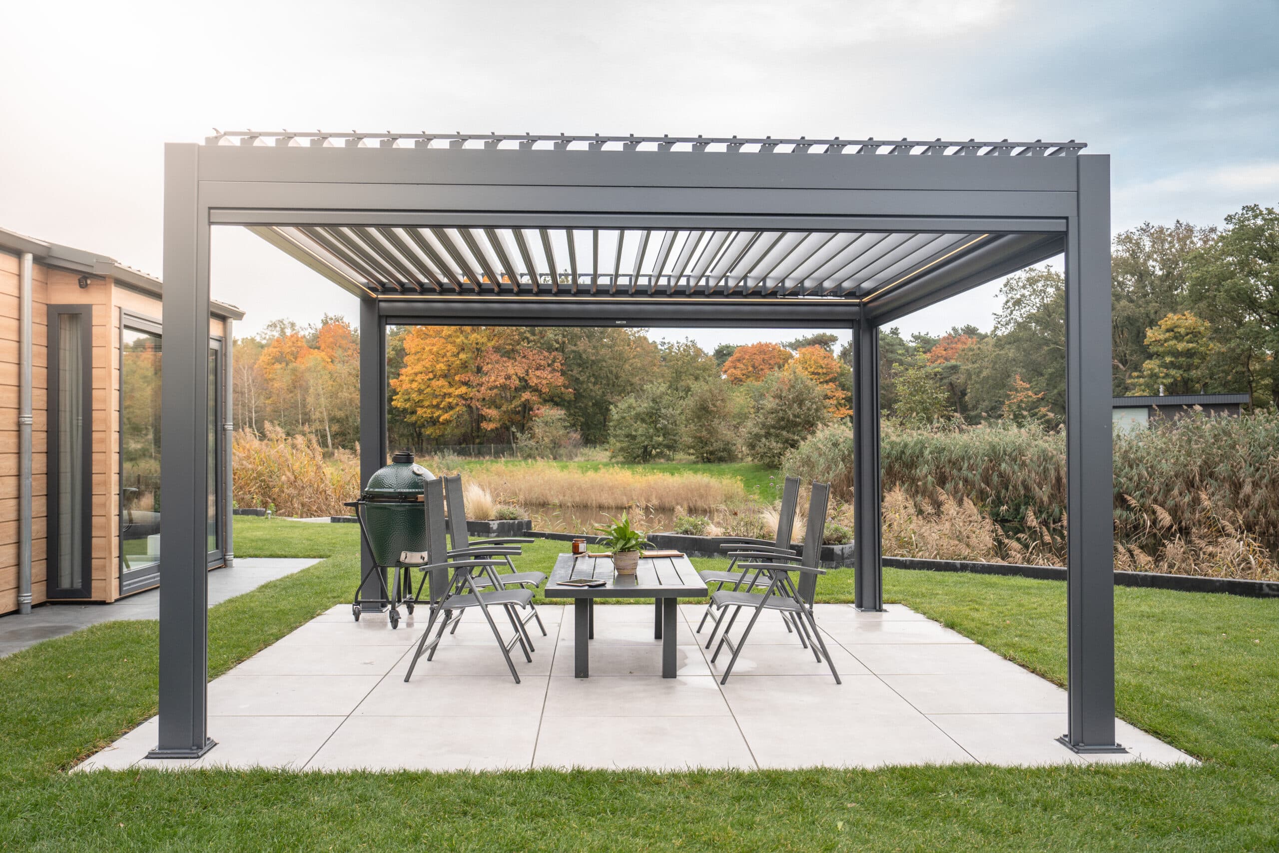 Eine moderne Außenterrasse mit einer Pergola aus Metall, für die eine Baugenehmigung für Terrassenüberdachung erforderlich ist, verfügt über acht Stühle und einen Tisch. An der Seite steht ein grüner Grill, umgeben von gepflegtem Gras und Bäumen, deren Herbstlaub unter einem teilweise bewölkten Himmel zu sehen ist.