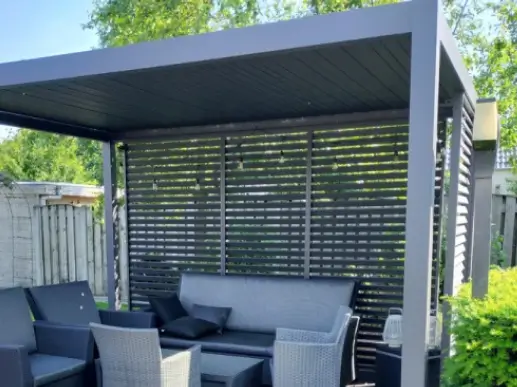 Eine moderne Außenterrasse mit einer eleganten grauen Pergola darüber. Drinnen stehen Korbstühle und ein gepolstertes Sofa um einen kleinen Tisch herum. Der Bereich ist von Laub umgeben und für die nötige Privatsphäre mit einer Holzlamellenwand abgetrennt.