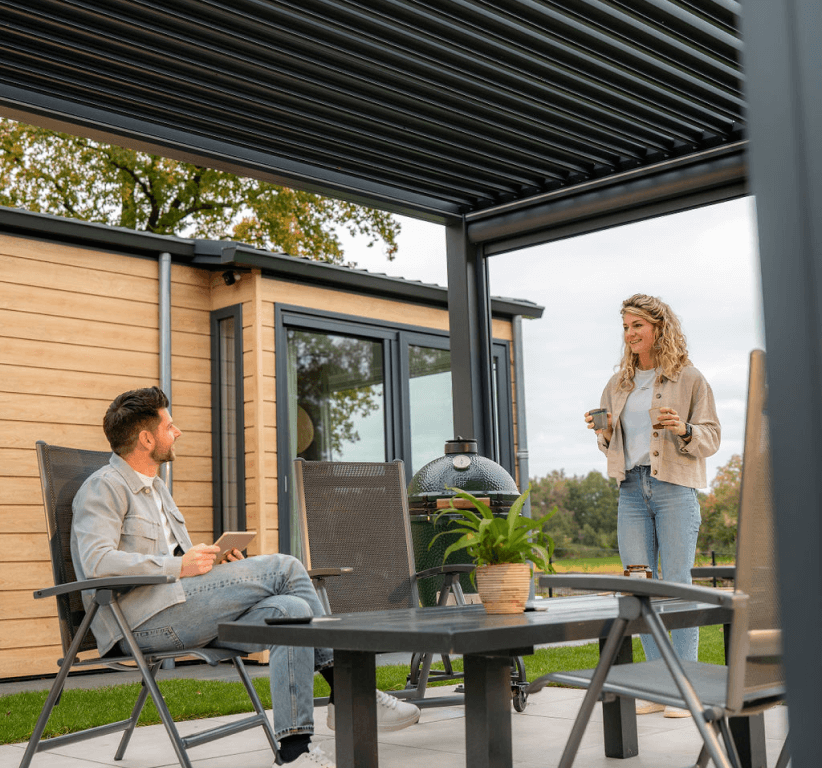 Ein Mann und eine Frau entspannen auf einer Terrasse unter einem Pergola-Garten. Der Mann sitzt mit einem Telefon da und unterhält sich ungezwungen mit der Frau, die neben ihm steht und an ihrer Tasse nippt. In der Nähe verleihen eine üppige Pflanze und ein großer Grill der Szene vor dem Hintergrund eines rustikalen Holzgebäudes Charme.