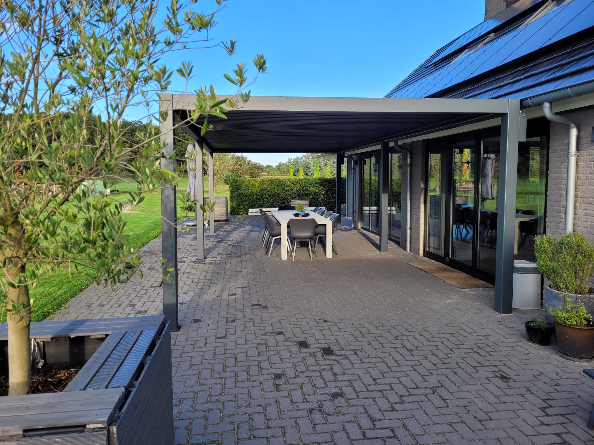 Eine moderne Außenterrasse mit Tisch und Stühlen unter einer nachhaltigen Aluminiumüberdachung. Der Bereich ist von Grün umgeben, darunter ein kleiner Baum in einem Pflanzkübel. Angrenzend an das Gebäude mit großen Glastüren genießt dieser Rückzugsort in Gaanderen einen klaren blauen Himmel.