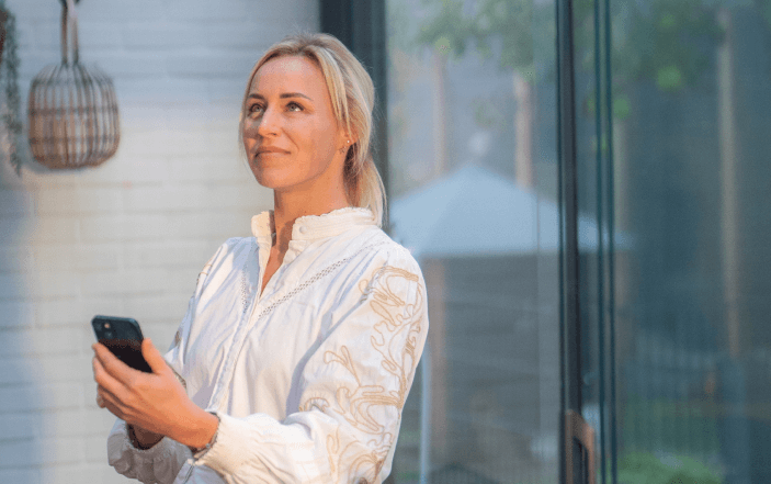 Eine Frau in einer weißen Bluse steht drinnen neben einem großen Fenster und hält ein Smartphone in der Hand. Sie lächelt und blickt nach oben, während natürliches Licht ihr Gesicht erhellt. Vor dem Fenster ist ein verschwommener Blick auf einen Garten zu sehen.