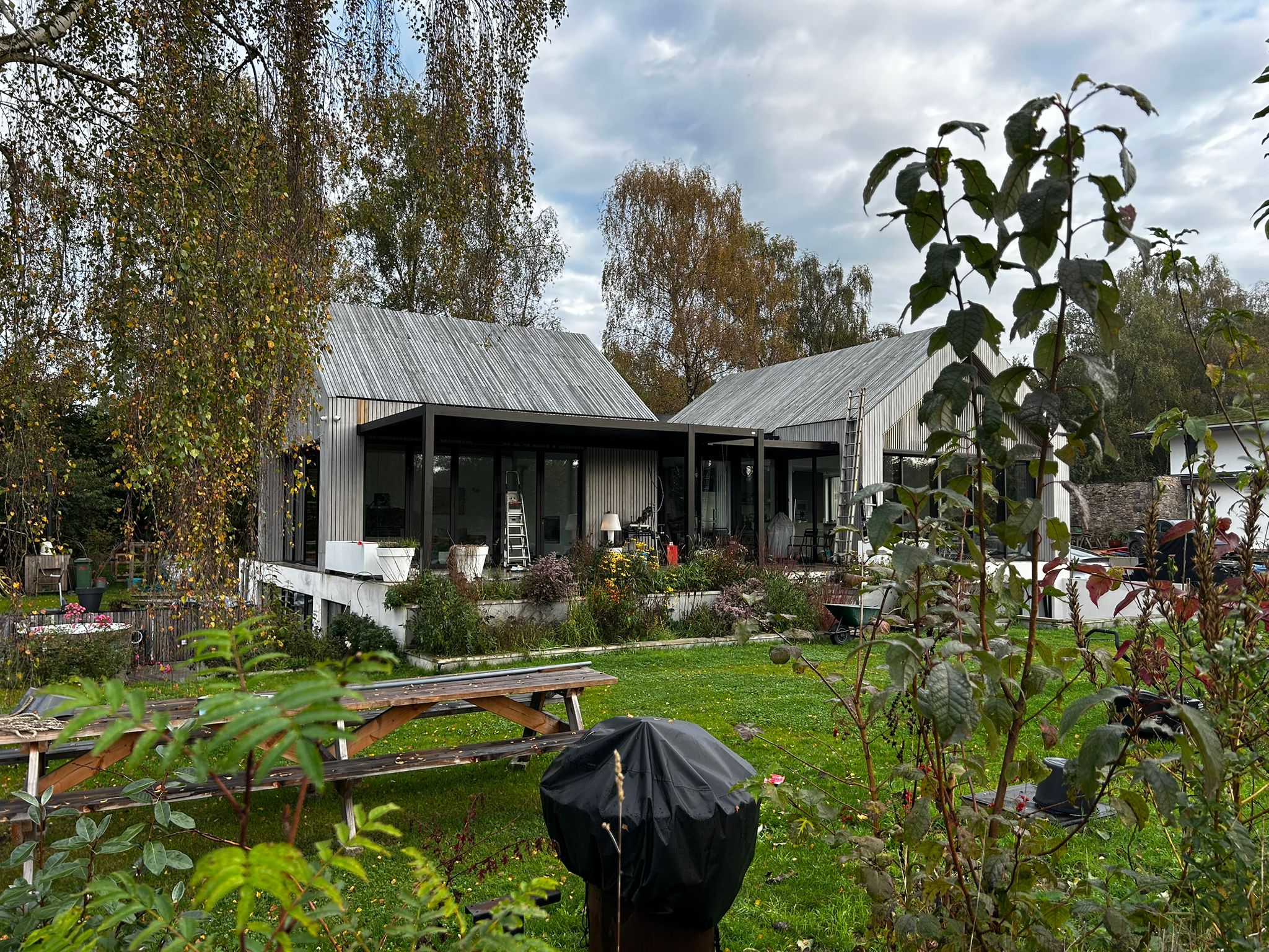 Ein modernes, L-förmiges Haus mit Metalldach in einem üppigen Garten. Das Haus verfügt über große Glasfenster und ist von viel Grün umgeben, darunter Bäume und Sträucher. Im Vordergrund sind ein hölzerner Picknicktisch und ein überdachter Grill zu sehen.