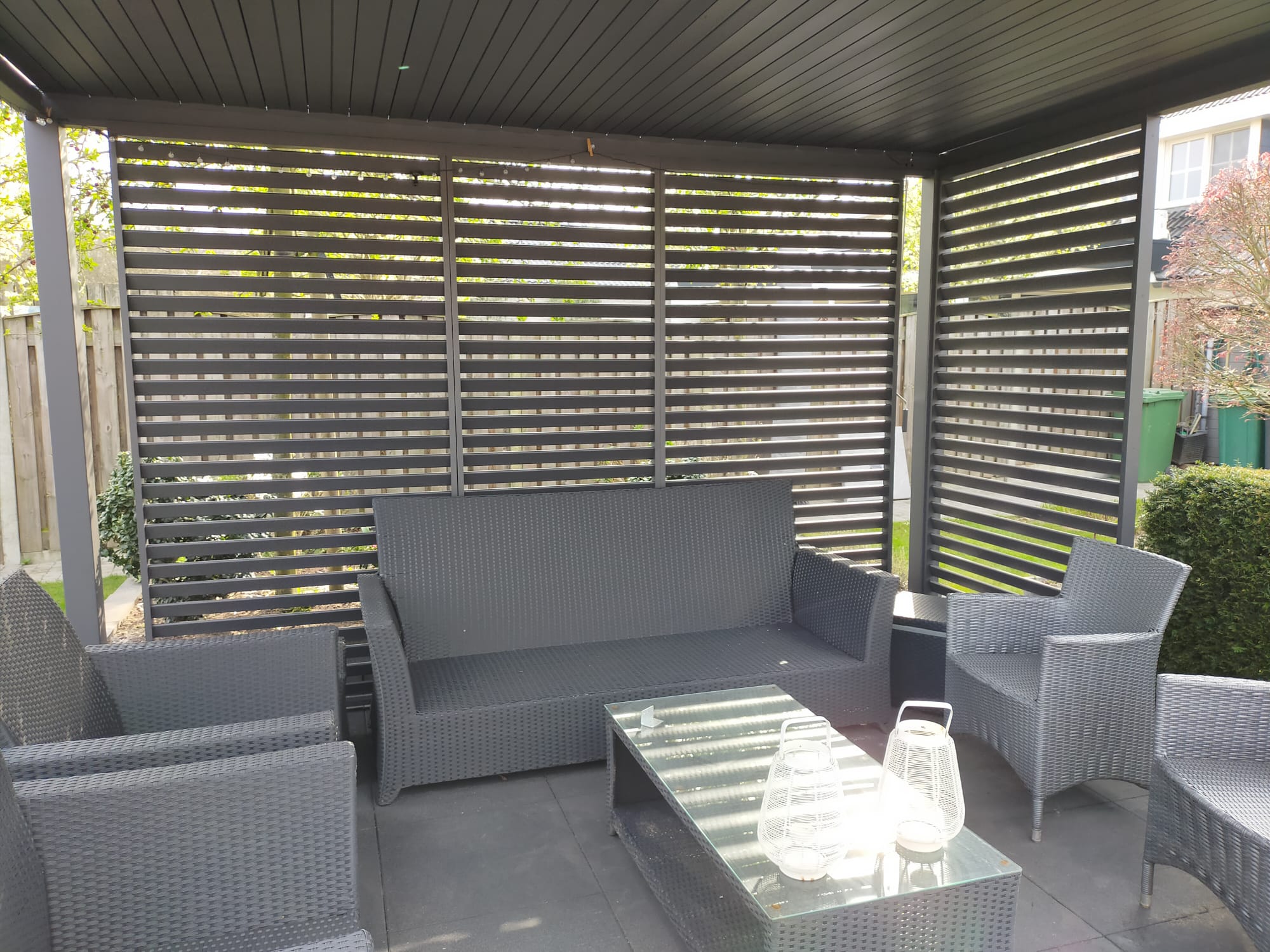 Außenterrasse mit dunkelgrauer Sitzgarnitur, darunter ein Sofa und Sessel, unter einer Pergola mit Lattenrost. Ein Couchtisch aus Glas hält dekorative Laternen. Der Bereich ist von Grünflächen und einem Holzzaun umgeben.