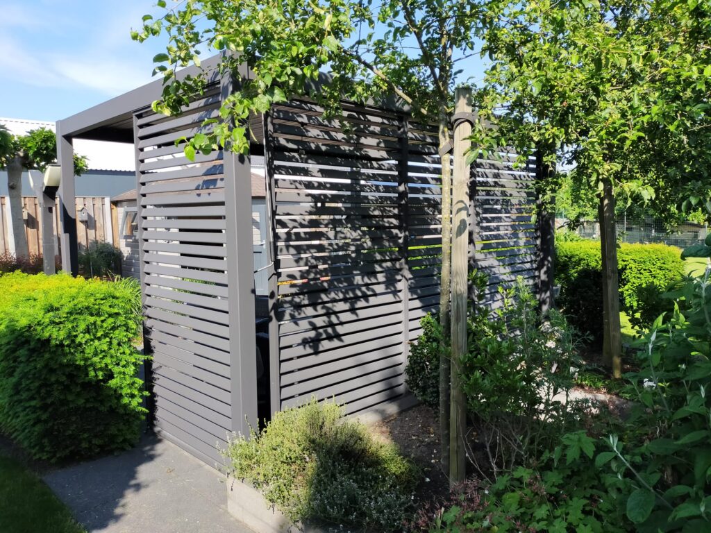 Eine moderne Pergola mit horizontalen Lamellenpaneelen ist von üppigen grünen Pflanzen und Sträuchern umgeben. Die Struktur spendet teilweise Schatten und im Hintergrund sind unter einem klaren blauen Himmel Bäume zu sehen.