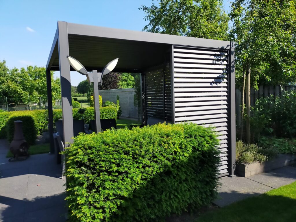 Eine moderne Pergola-Struktur mit horizontalen Lamellen ist von üppigen grünen Büschen umgeben. Zwei minimalistische Laternenpfähle stehen in der Nähe. Die Umgebung umfasst Bäume und einen Weg mit einer ruhigen, sonnigen Außenatmosphäre.