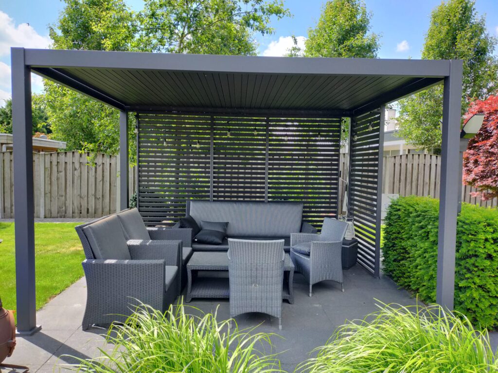 Ein moderner Sitzbereich im Freien mit einer Pergola aus Metall. Er verfügt über ein schwarzes Sofa, mehrere geflochtene Stühle und einen niedrigen Tisch auf einer Steinterrasse. Der Raum ist von üppigem Grün und einem Holzzaun umgeben. Der Himmel ist klar mit ein paar Wolken.