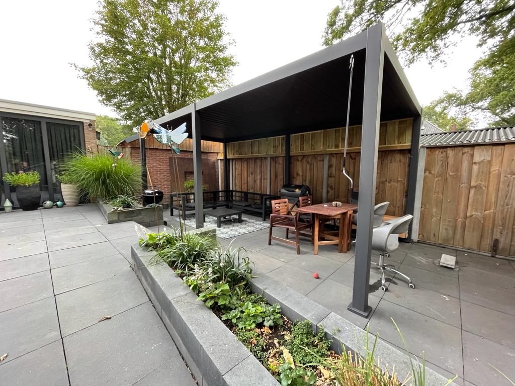 Eine Hinterhofterrasse mit einem modernen, seitlich offenen Pavillon mit dunklem Metallrahmen und Lamellenüberdachung. Im Inneren gibt es einen Sitzbereich mit einer Bank und einem Holztisch. Topfpflanzen umgeben die graue Steinterrasse und ein großer Baum spendet teilweise Schatten.