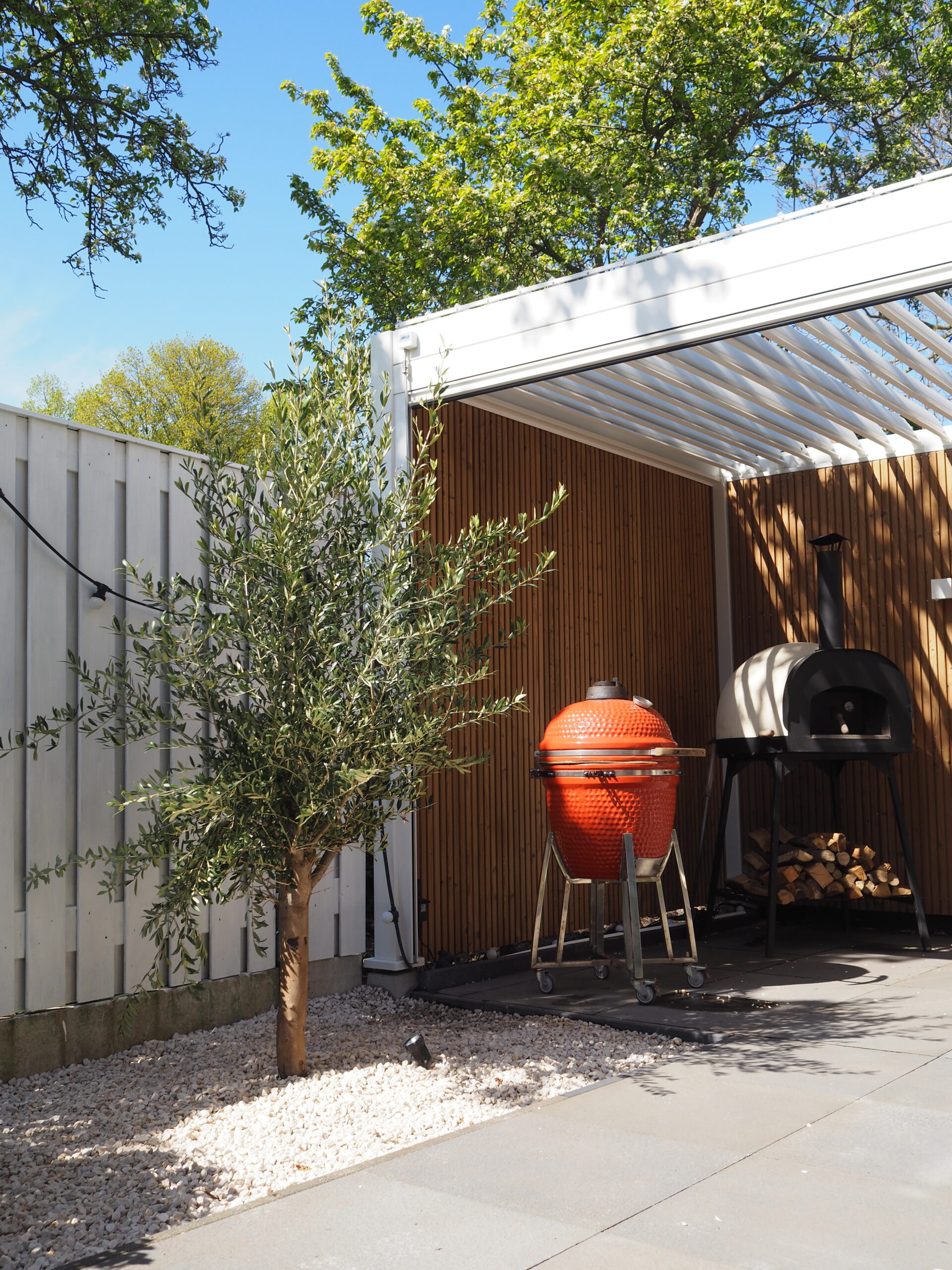 Grillplatz im Freien mit einem roten Keramik-Smokergrill und einem schwarzen Pizzaofen unter einer weißen Lamellenüberdachung. Ein Olivenbaum steht in der Nähe auf einem mit Kieselsteinen bedeckten Platz, der von einem Holzzaun umgeben ist. Neben dem Pizzaofen ist ein Stapel Brennholz zu sehen, der diese charmante Kundengeschichte noch verstärkt.