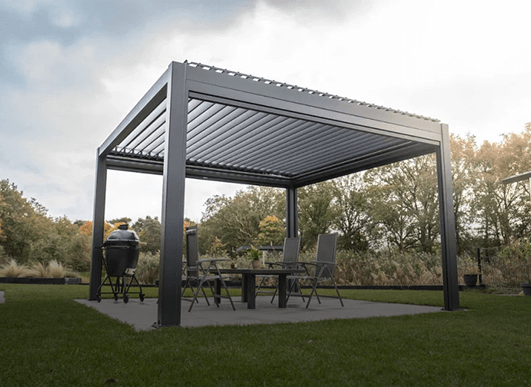 Eine moderne Metallpergola mit Lamellendach steht auf einer Terrasse in einem grasbewachsenen Hinterhof und bedeckt einen Tisch mit vier Stühlen und einen Grill. Im Hintergrund sind Bäume und Sträucher unter einem teilweise bewölkten Himmel zu sehen.