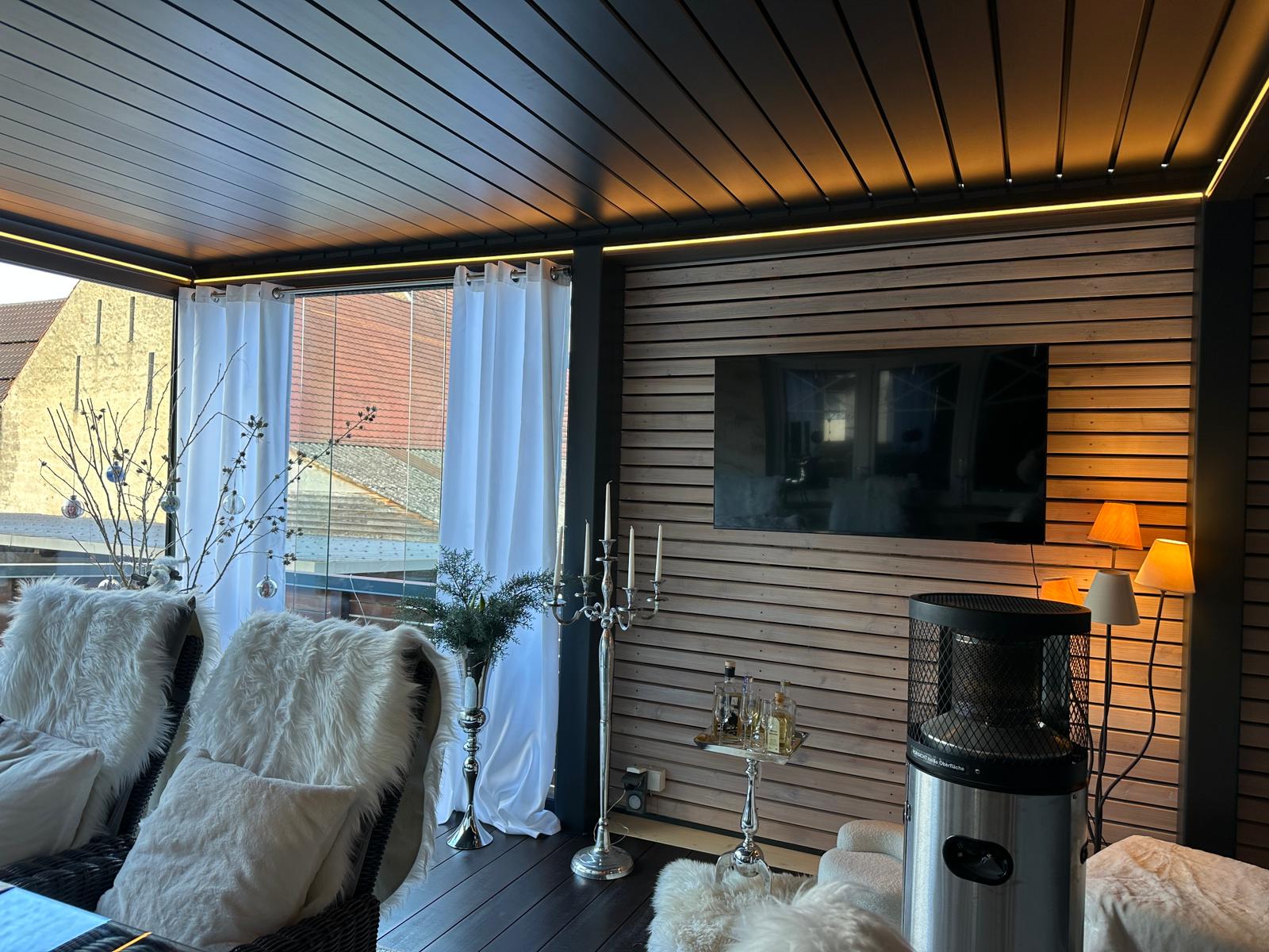 Ein gemütlicher Sitzbereich im Innenbereich bietet Korbstühle mit weißen Felldecken und einen Fernseher an der Holzwand. Ein moderner Heizstrahler wärmt den Raum, während große Fenster mit weißen Vorhängen und hellem Dekor den Blick nach draußen unter die charmante Lamellenüberdachung ermöglichen.