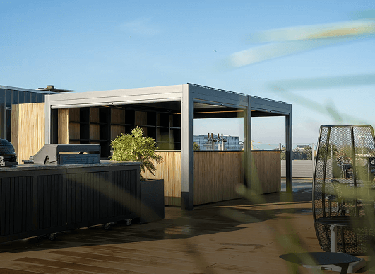 Eine moderne Dachterrasse mit Grill, Außenküche, Sitzbereich und Pergola-Struktur unter einem klaren blauen Himmel mit etwas sichtbarem Grün.