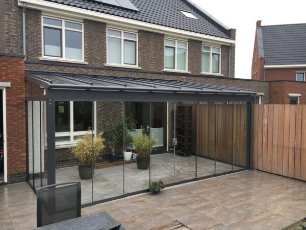 Ein modernes Haus mit einer verglasten Terrasse, ausgestattet mit der Terrassenüberdachung nach Maß, einem Metallrahmendach, Topfpflanzen, großen Fliesenböden und einem Zaun aus Holzpaneelen.