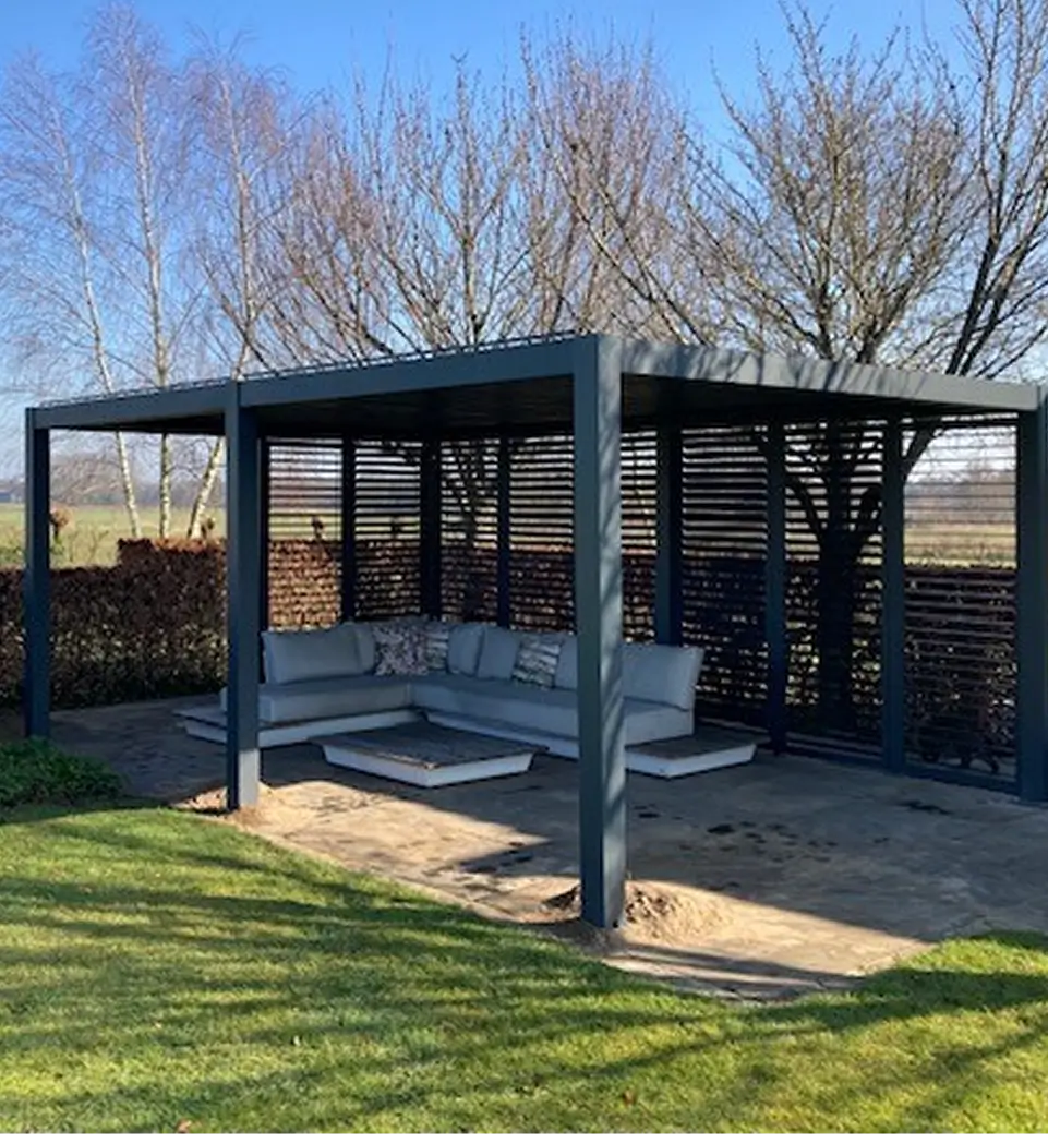 Eine moderne Pergola mit dunklem Metallrahmen und Lattenrosten schützt ein graues, gepolstertes L-förmiges Sofa und einen Couchtisch, die auf einer Steinterrasse in einem grasbewachsenen Garten mit kahlen Bäumen im Hintergrund stehen.