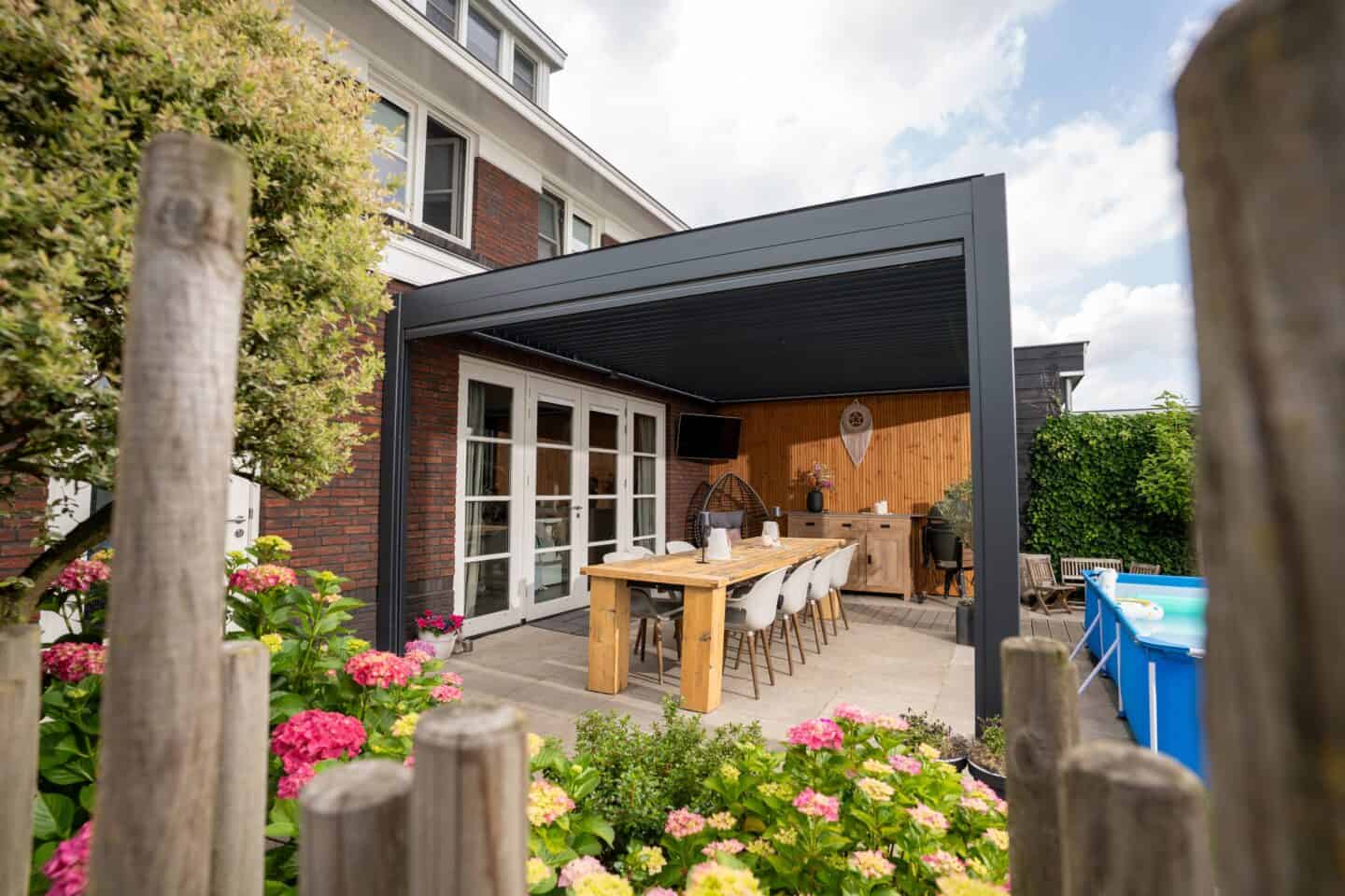 Am Haus montierte Lamellenüberdachung mit Esstisch in einem blühenden Garten.